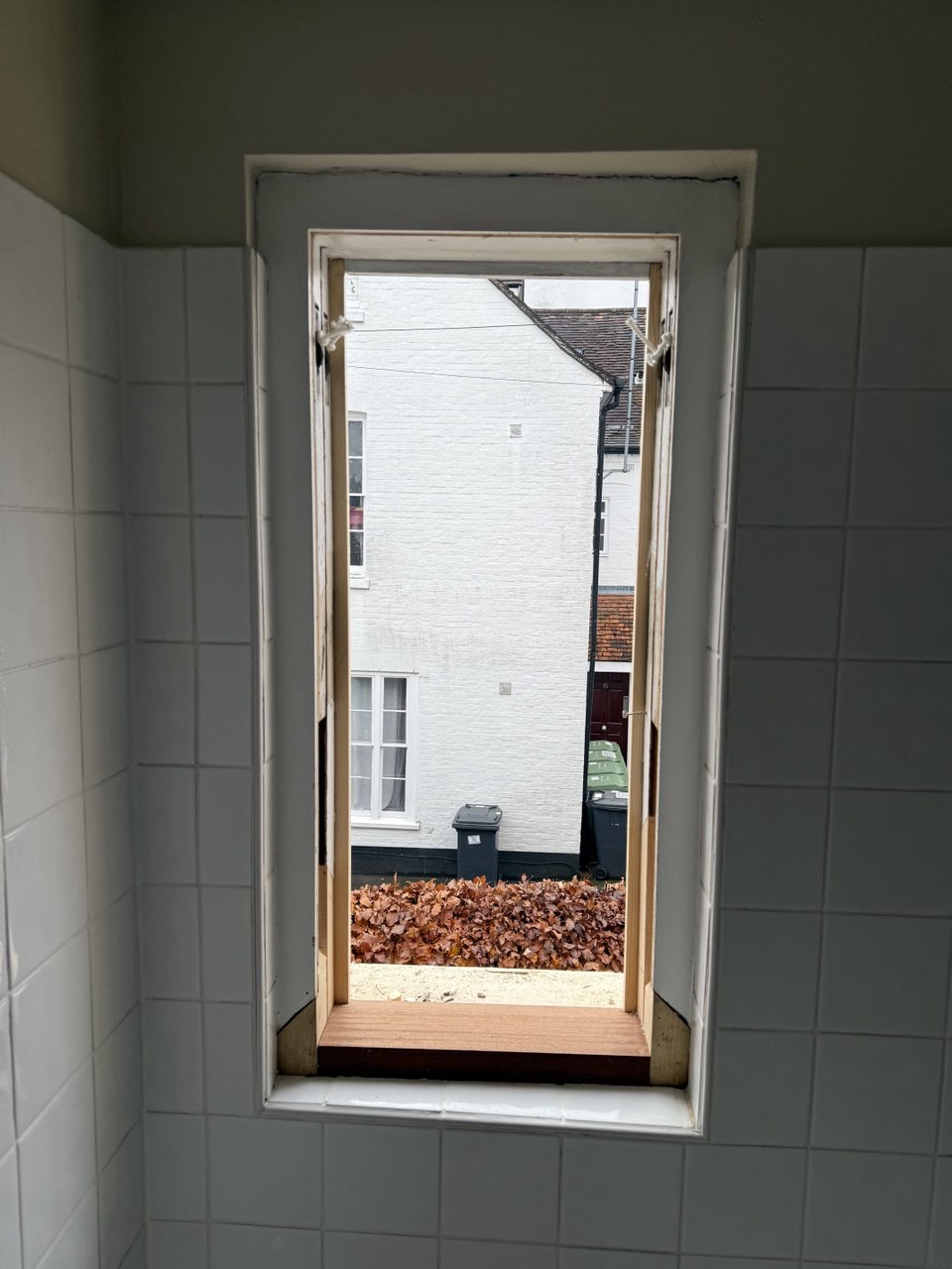 A small open sash window in a tiled wall reveals a view of a white building, a rubbish bin, and brown leaves outside—reminiscent of the classic architecture found in Cambridge.